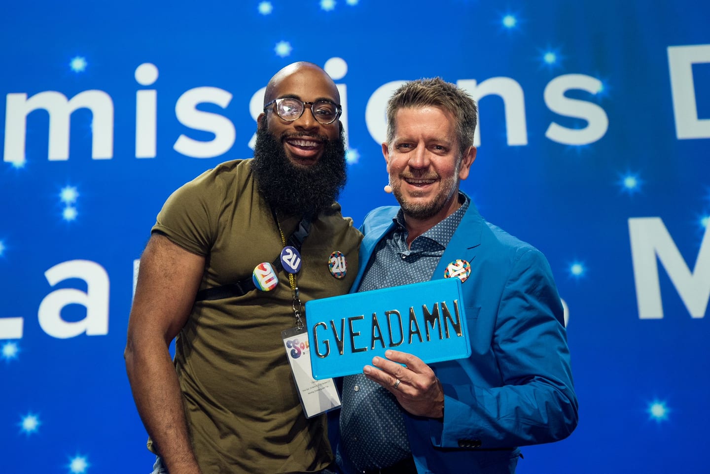 Two employees on stage holding a GVEADAMN licence plate award against a blue starfield screen