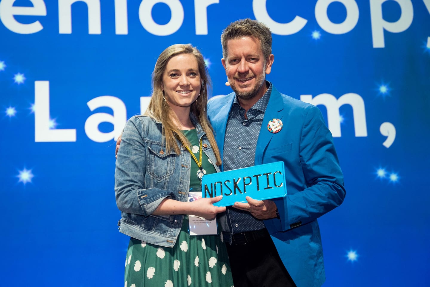 Two employees on stage holding a blue NOSKPTIC licence plate award in front of a bright screen