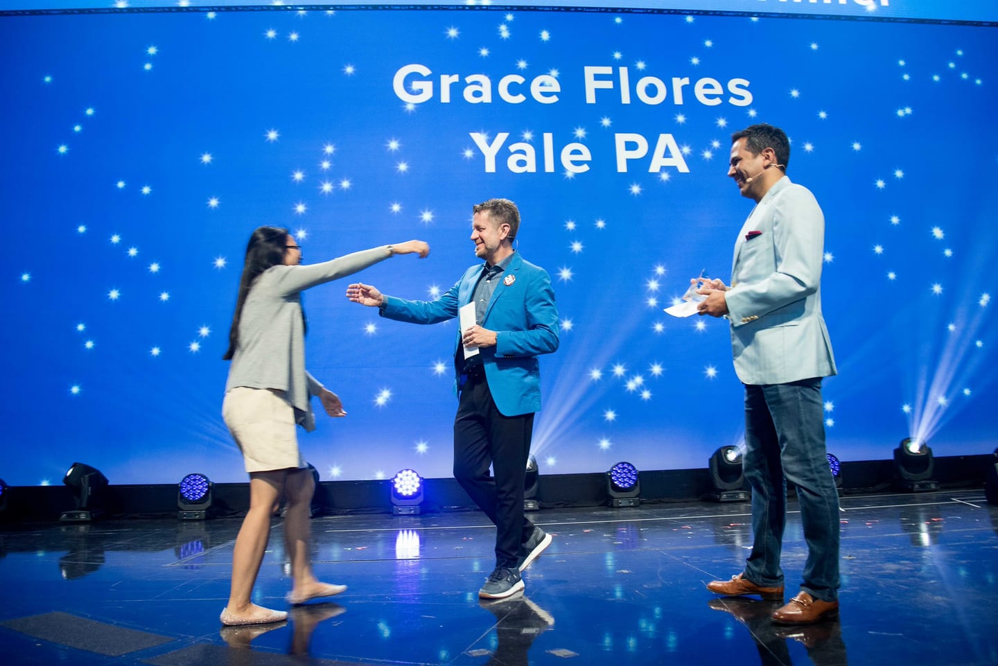 An award recipient walking on to the Colosseum stage as two hosts applaud beneath a blue screen