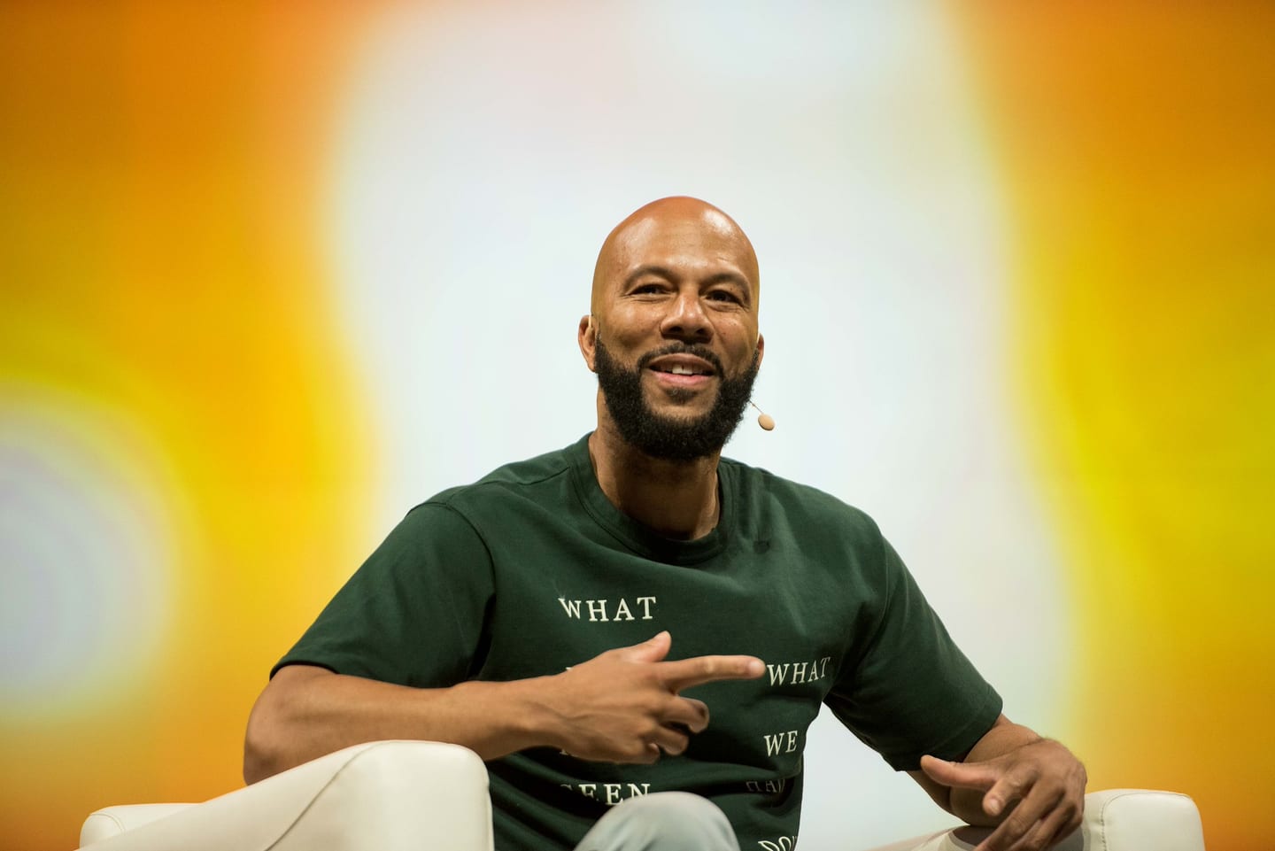 Common smiling mid-conversation in a white armchair against a warm orange stage screen