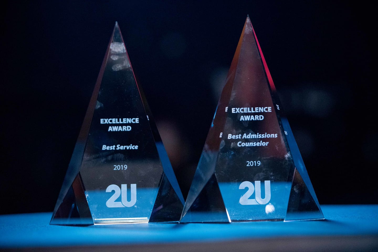 Two clear 2U Excellence Award trophies lit blue on a table, engraved Best Service and Best Admissions Counselor