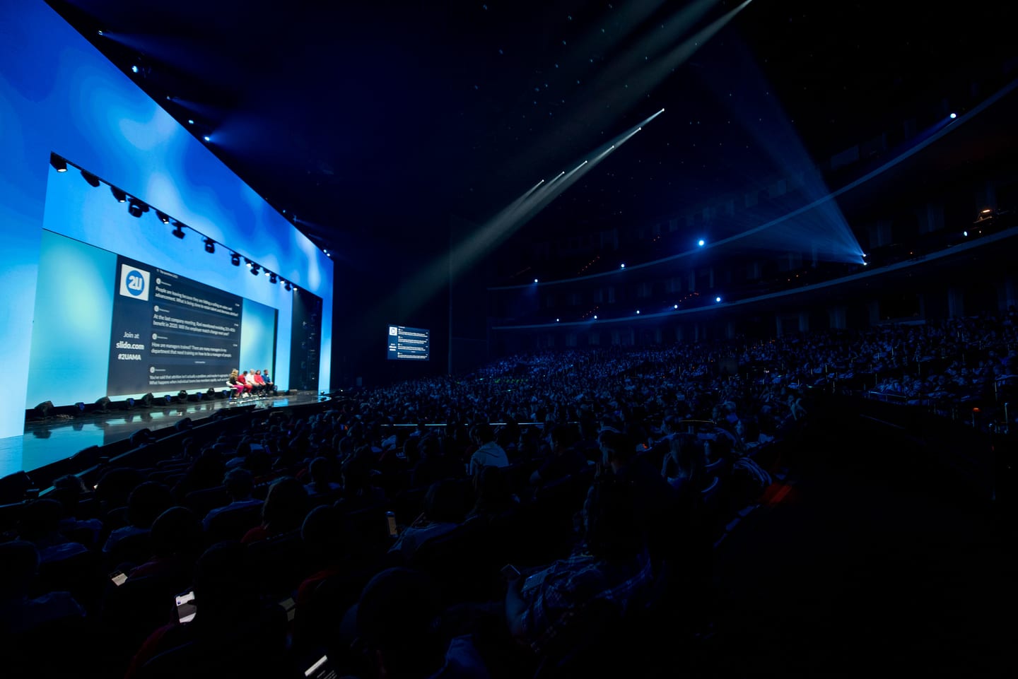 A wide view of the Colosseum during a general session, blue stage light spilling over a full seated audience