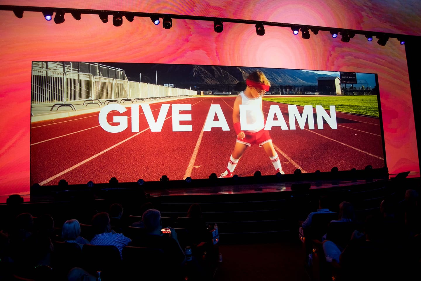 The words GIVE A DAMN across the full Colosseum screen above a runner on a track, seen over the audience