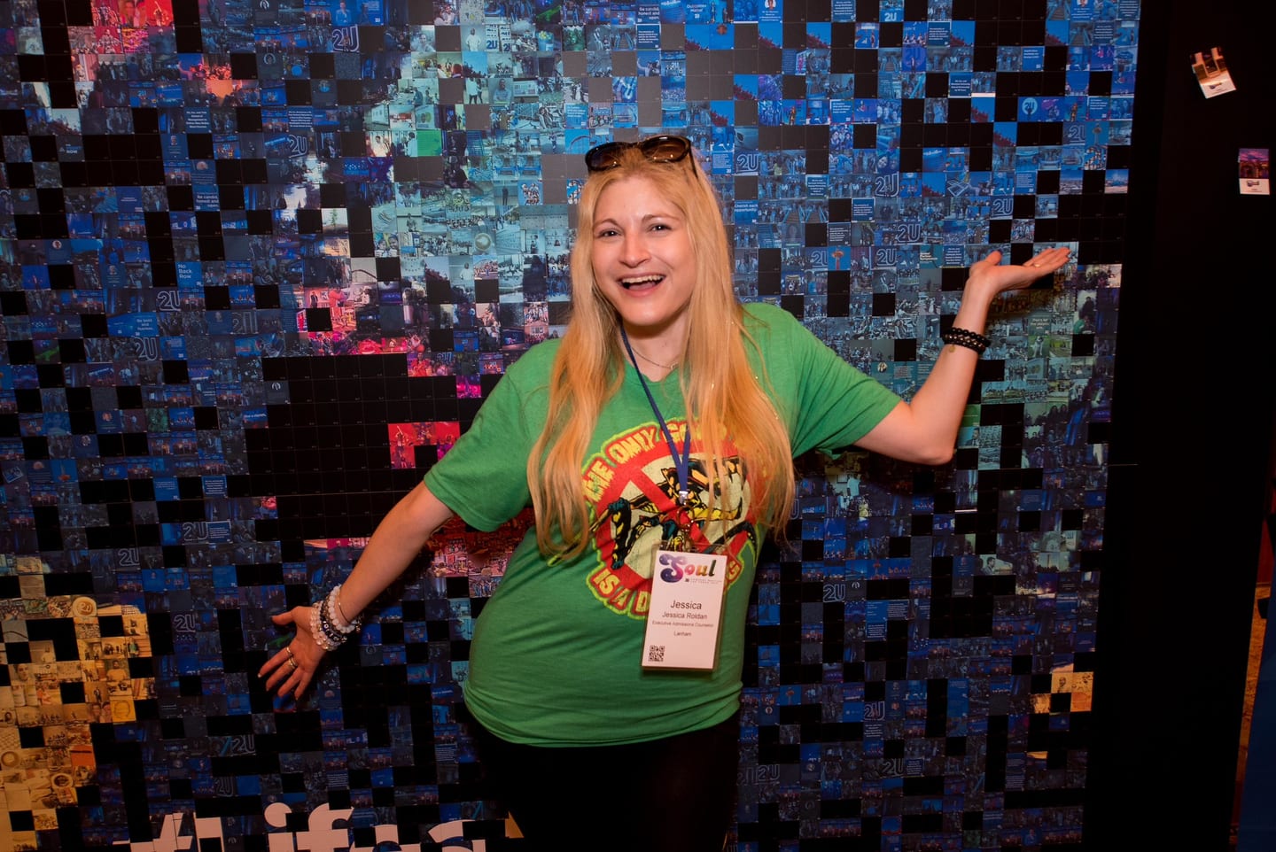 An employee in a green t-shirt gesturing in front of a large tiled photo-mosaic wall