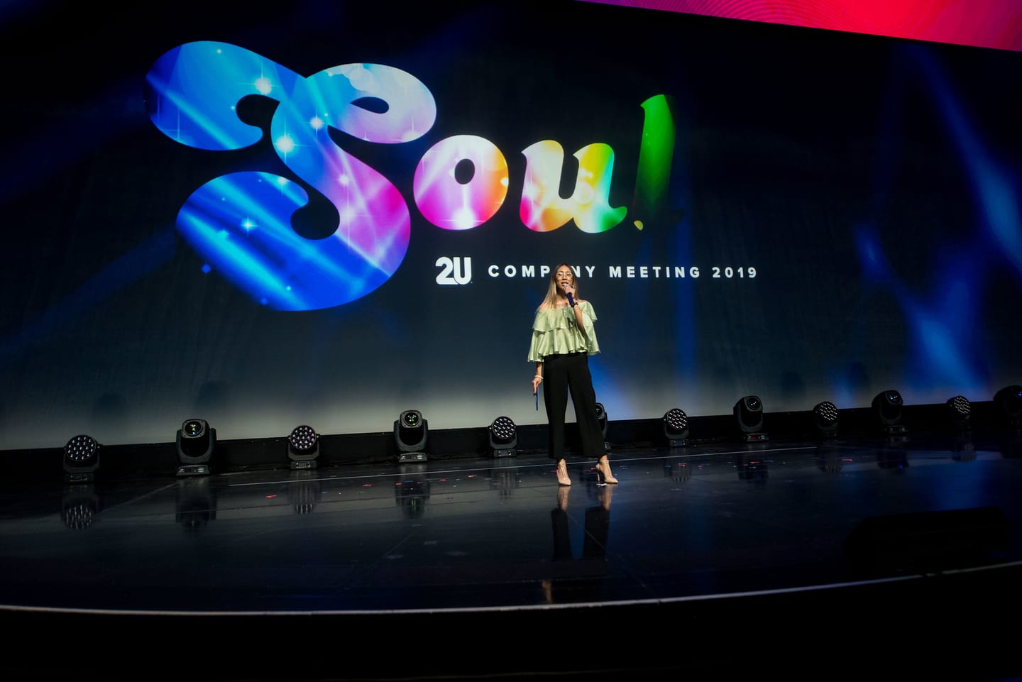 A presenter standing centre stage at the Colosseum beneath the SOUL 2U Company Meeting 2019 screen