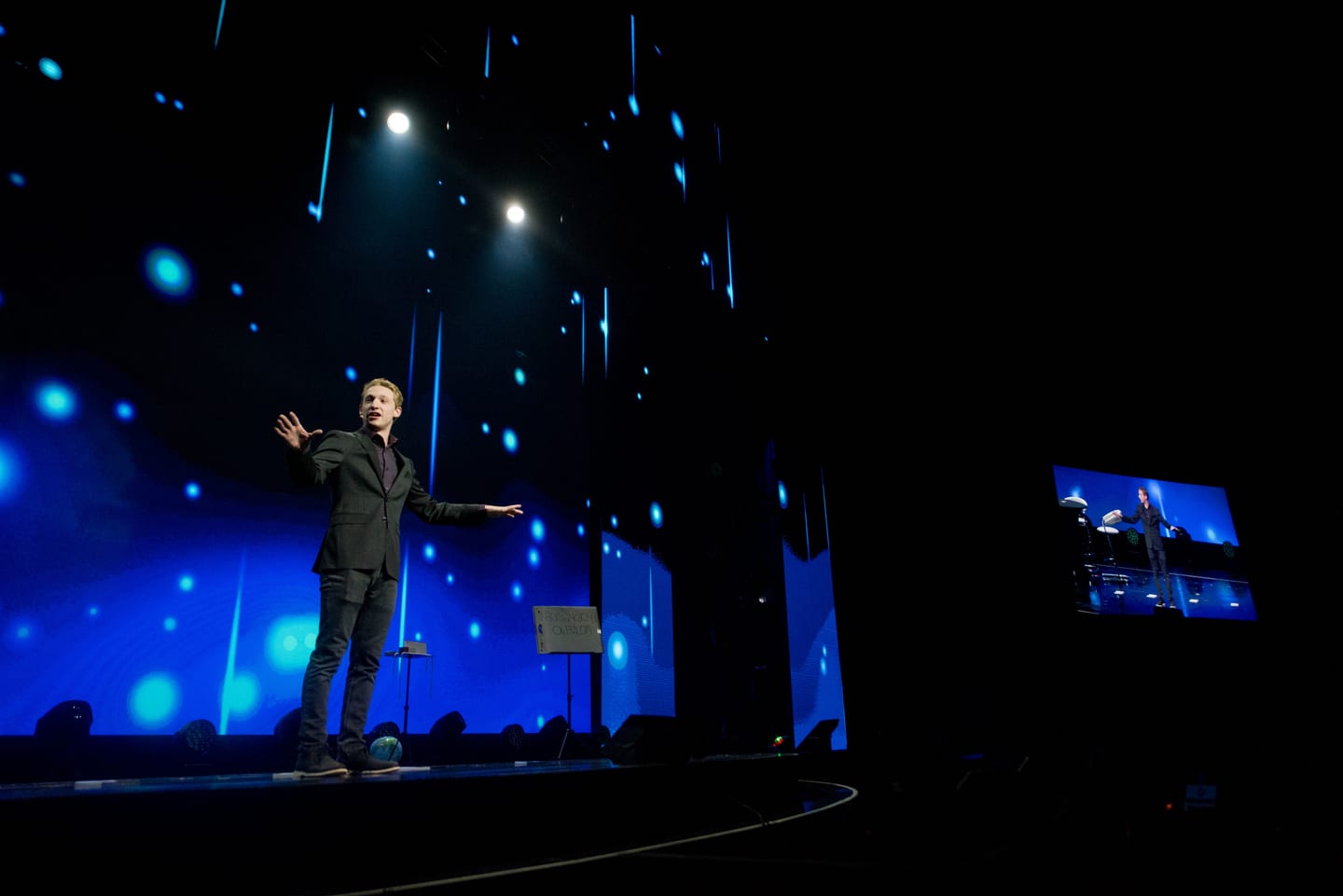 A lone speaker on the wide Colosseum stage lit blue with the audience in darkness