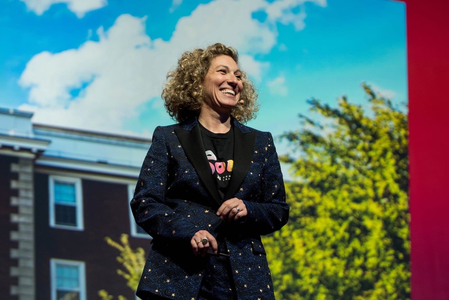 A speaker smiling on stage in front of a screen showing a brick campus building and trees