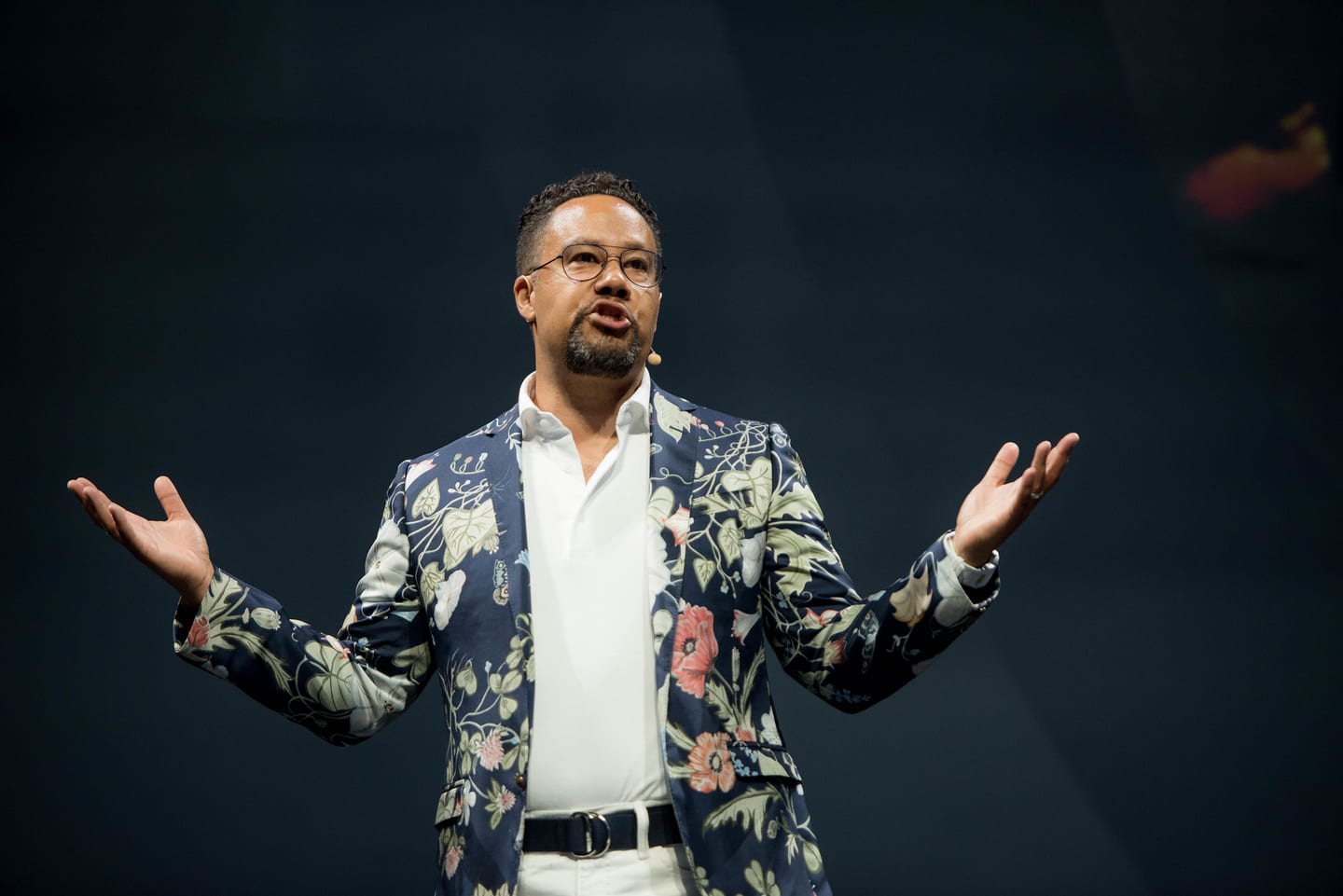 A speaker in a floral jacket presenting with both arms open on the Colosseum stage