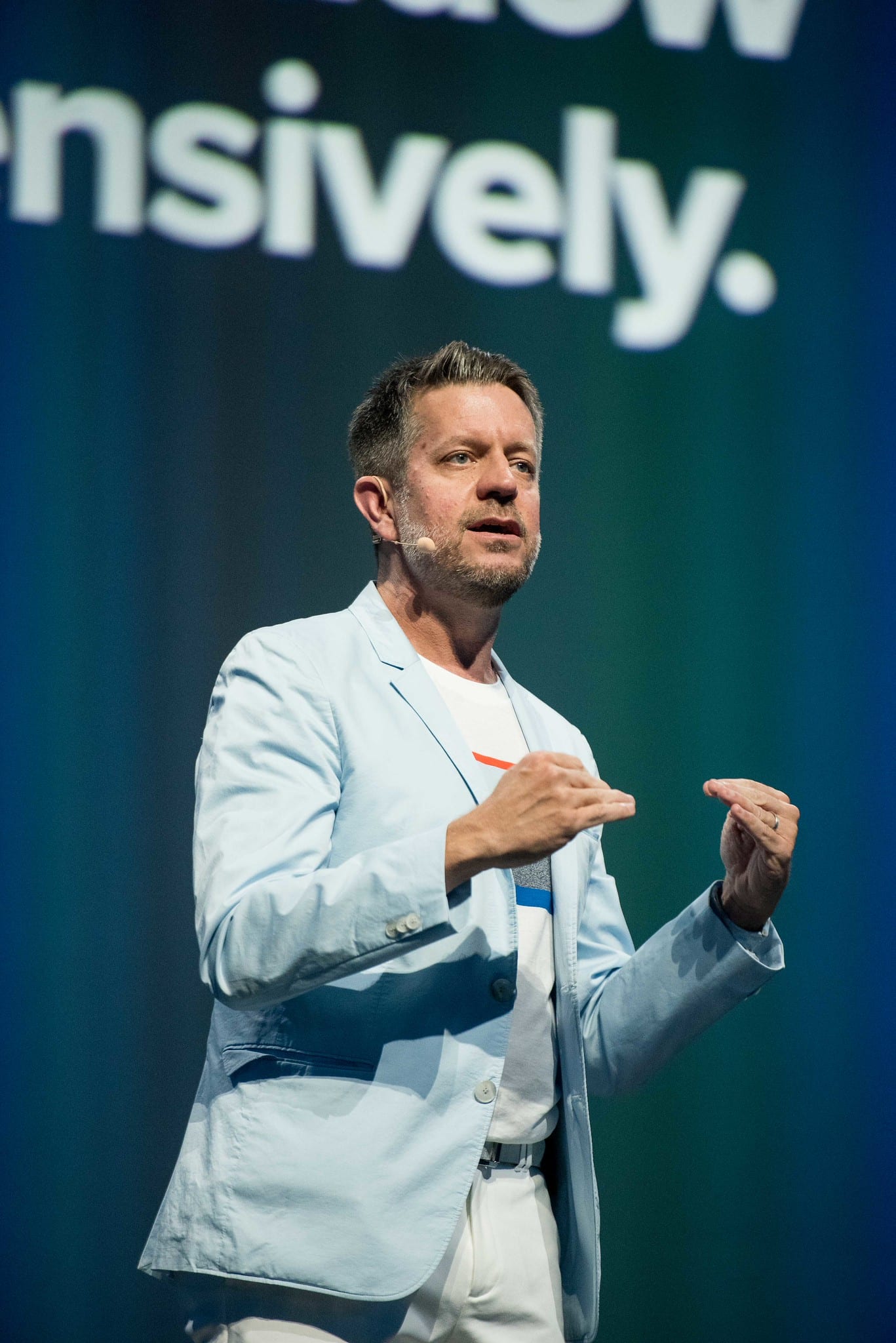 A speaker in a pale blue jacket gesturing in front of a screen reading grow intensively