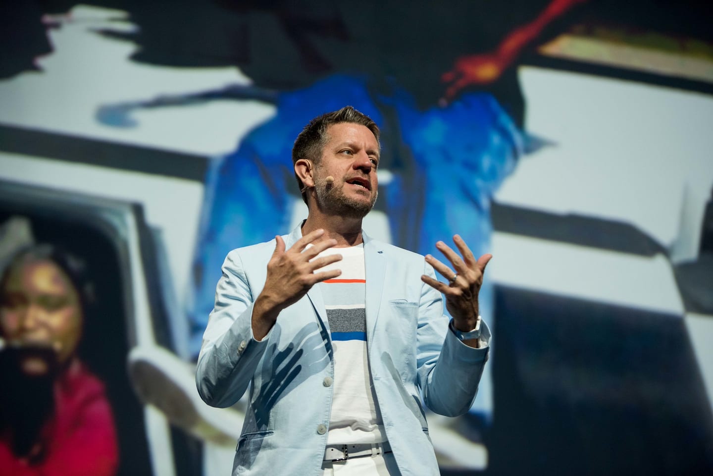 A speaker in a pale jacket gesturing mid-sentence on the Colosseum stage