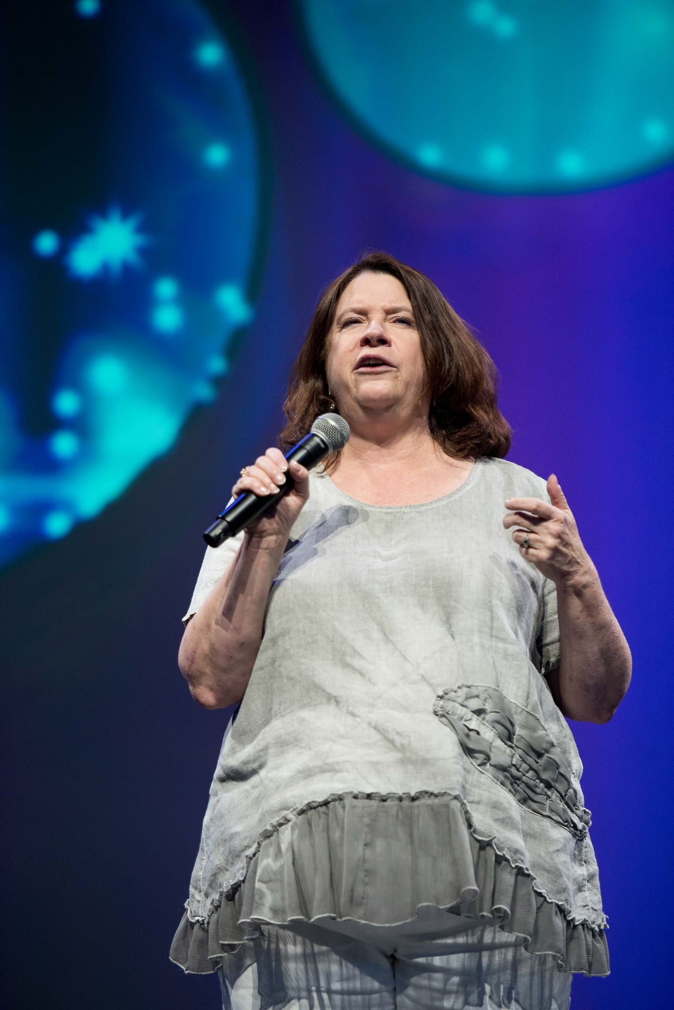 A speaker holding a handheld microphone on stage against a blue and purple screen