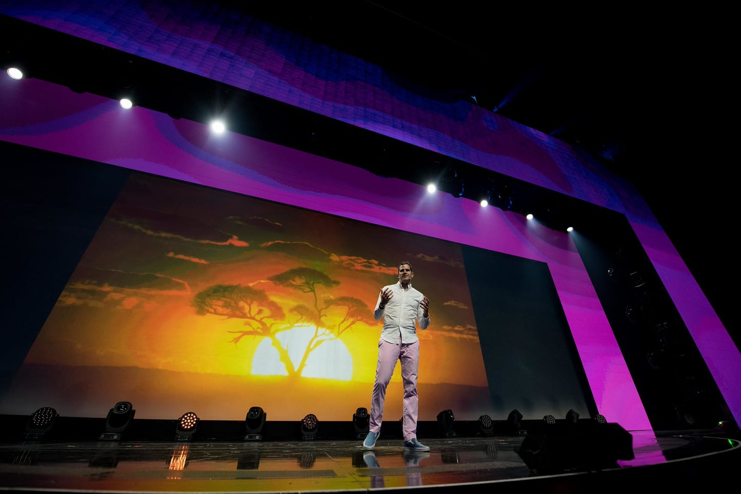A speaker crossing the Colosseum stage in front of a wide screen showing a savanna sunset