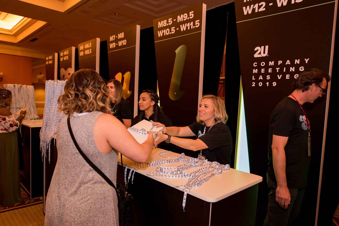 Employees at a swag table under signs listing shoe sizes at the 2U company meeting