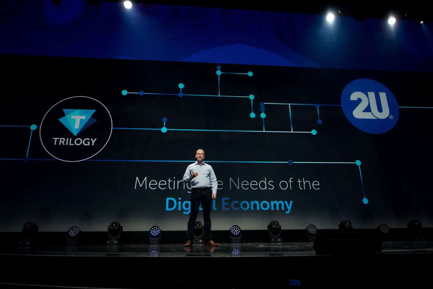A speaker on stage beside a screen reading Meeting the Needs of the Digital Economy with the Trilogy and 2U logos