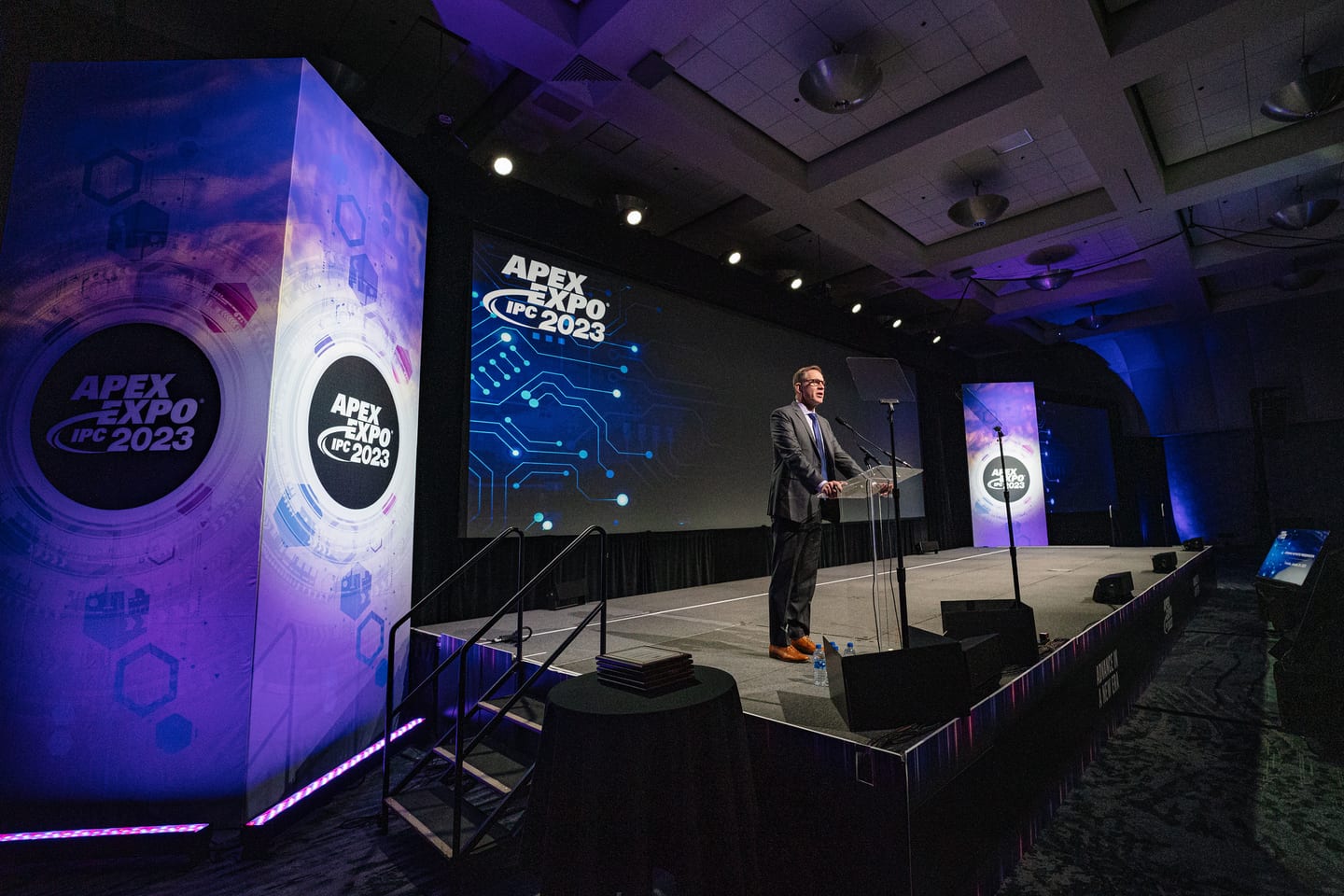 A speaker at the podium on a purple-lit main stage under the IPC APEX EXPO 2023 branding