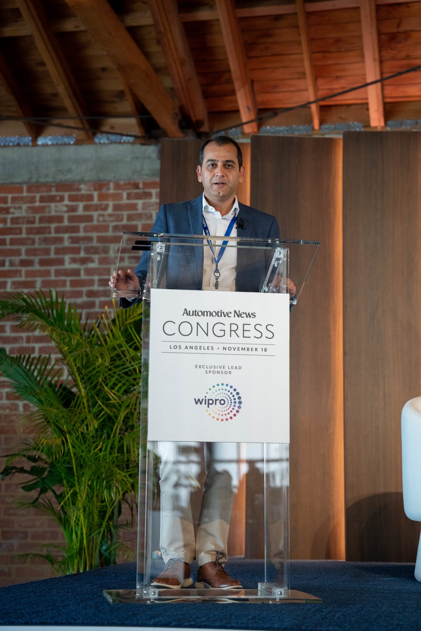 A speaker at the lectern seen wide, with the empty stage chairs beside them at the Automotive News Congress: Los Angeles, City Market Social House