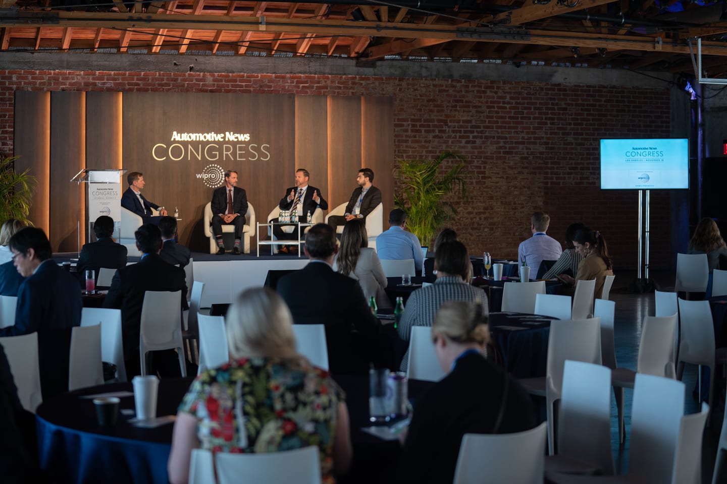 A wide view of the room with a panel on stage and attendees seated at round tables under the timber trusses at the Automotive News Congress: Los Angeles, City Market Social House