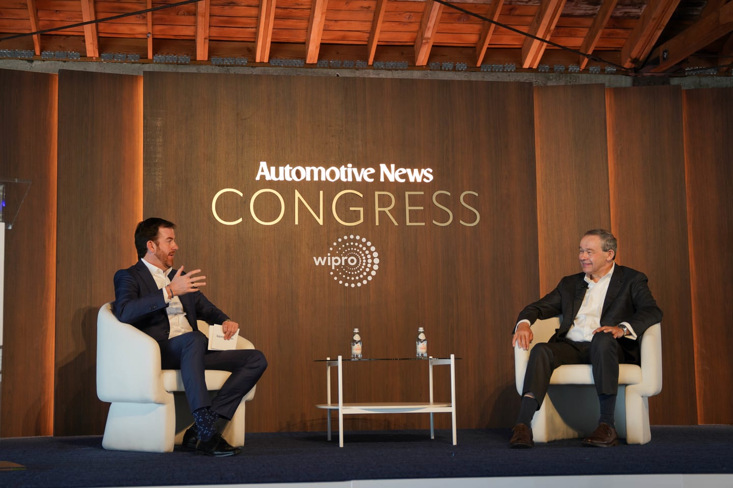 Two executives in a fireside conversation on stage beneath the illuminated Automotive News Congress wall and Wipro lead-sponsor mark at the Automotive News Congress: Los Angeles, City Market Social House