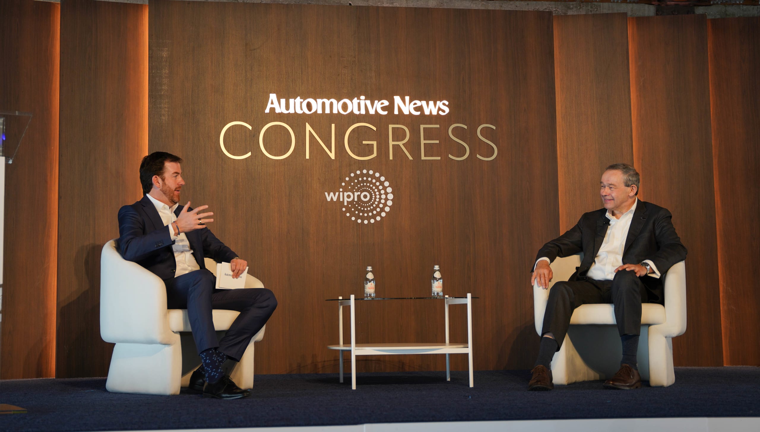 Two executives in a fireside conversation on stage beneath the illuminated Automotive News Congress wall and Wipro lead-sponsor mark at the Automotive News Congress: Los Angeles, City Market Social House