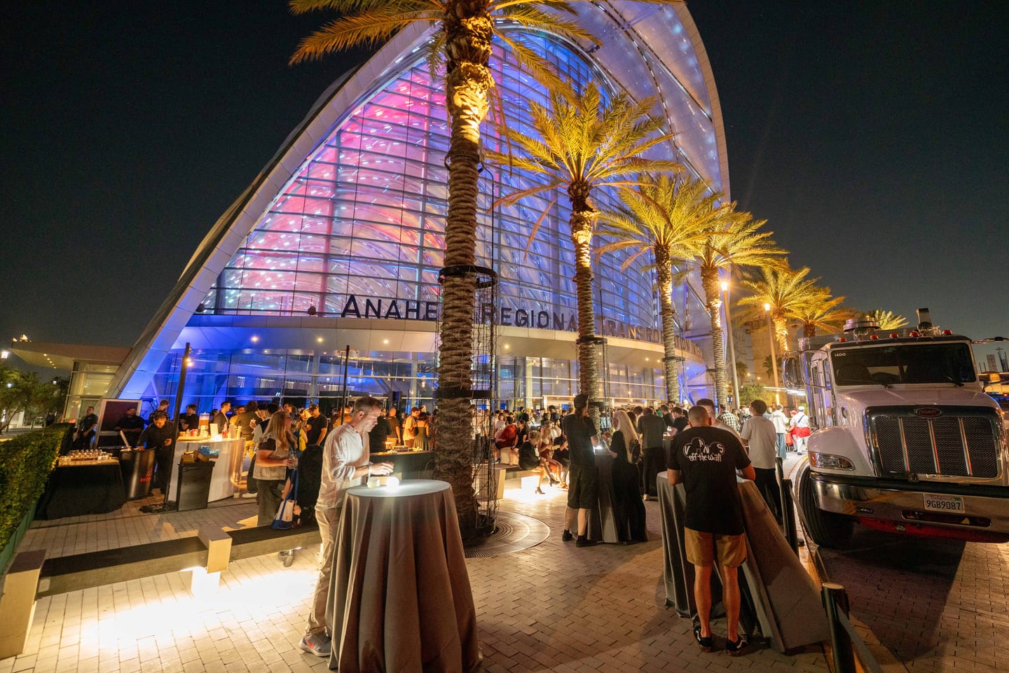 Guests at cocktail tables on the patio outside ARTIC at night, with the lit arched facade and palm trees behind them