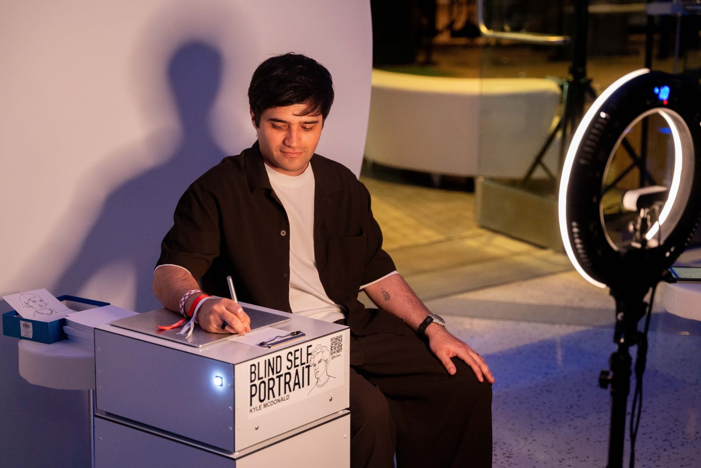 A guest drawing on a Blind Self Portrait machine labeled Kyle McDonald, beside a ring light