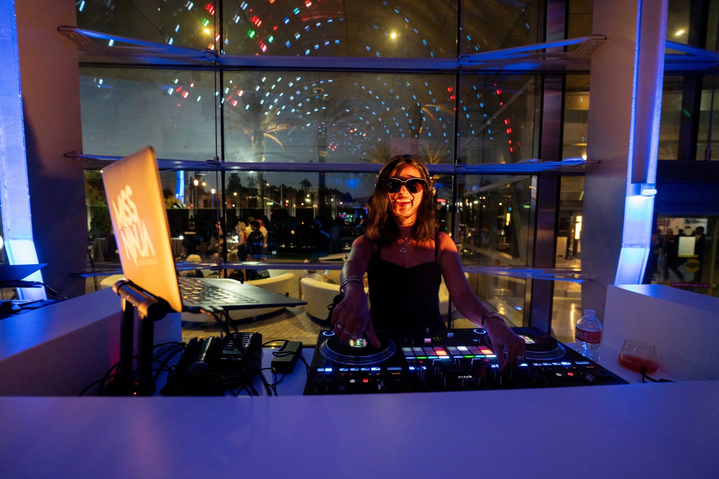A DJ at the decks with a Miss Ninja laptop, in front of the glass wall of ARTIC at night