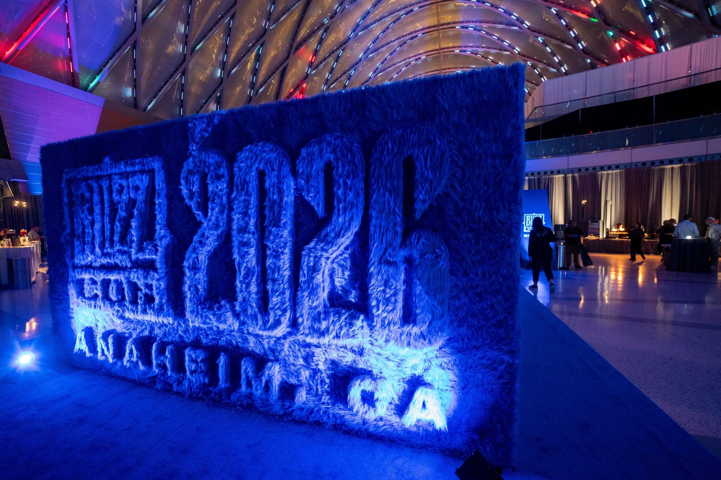 An eight-foot blue shag wall spelling BlizzCon 2026 Anaheim, CA, lit blue under the arched roof of ARTIC in Anaheim