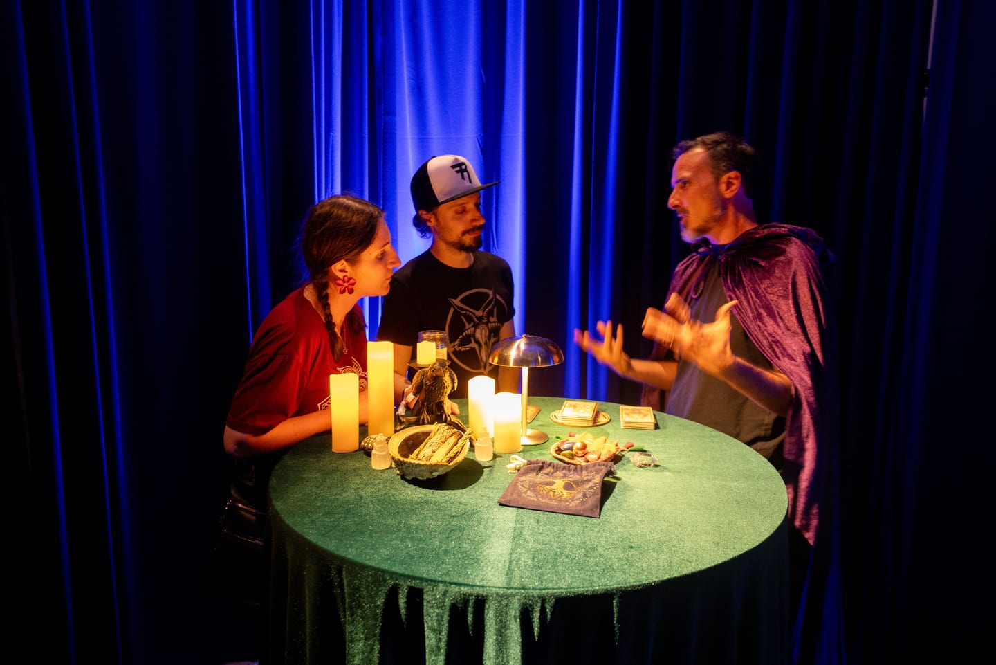 A tarot reader in a purple cape talking with two guests at a candlelit green table in front of blue curtains