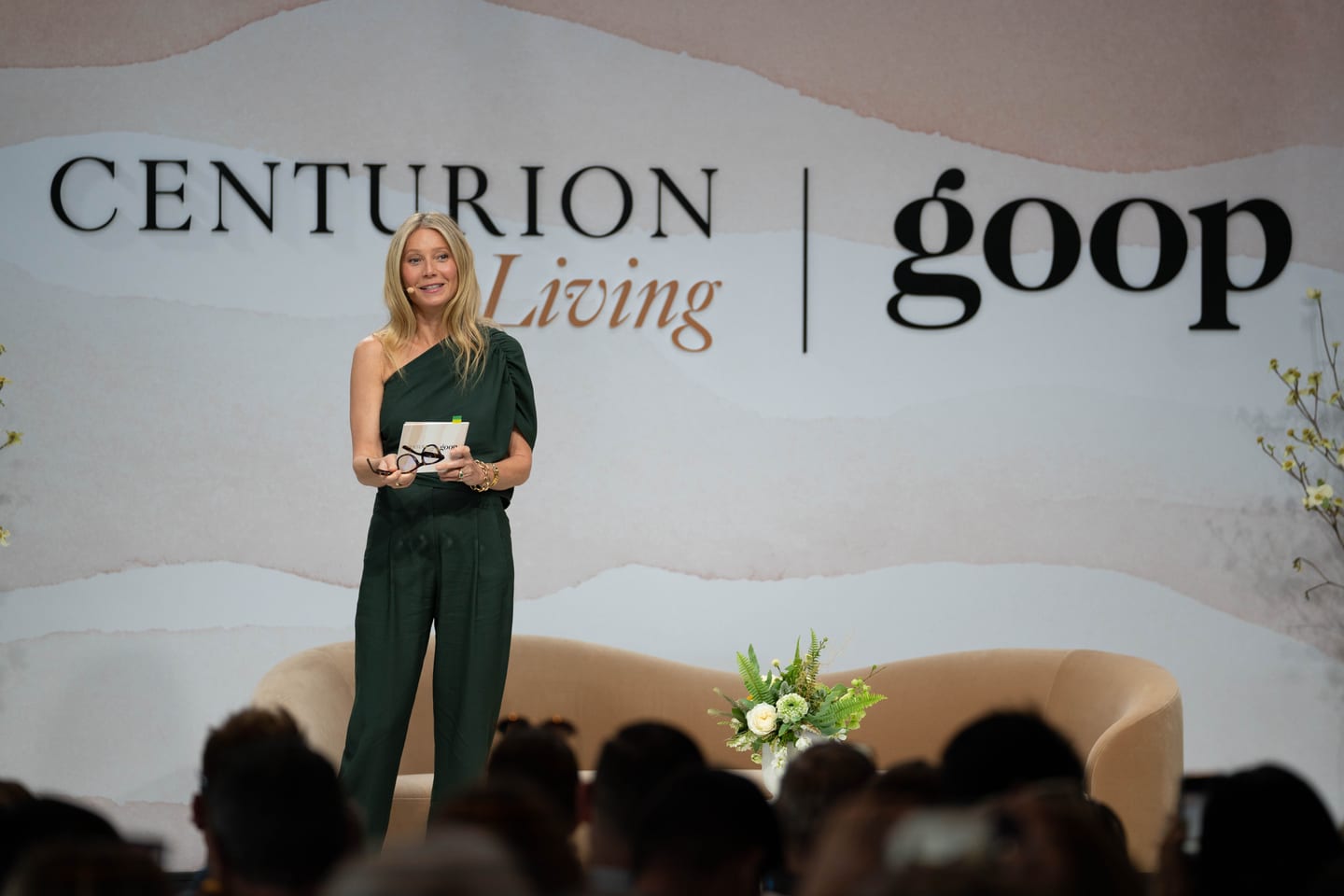 Gwyneth Paltrow in a dark green outfit speaking on stage with a headset microphone in front of a large CENTURION Living and goop backdrop, a curved sofa and florals behind her and the audience silhouetted in front