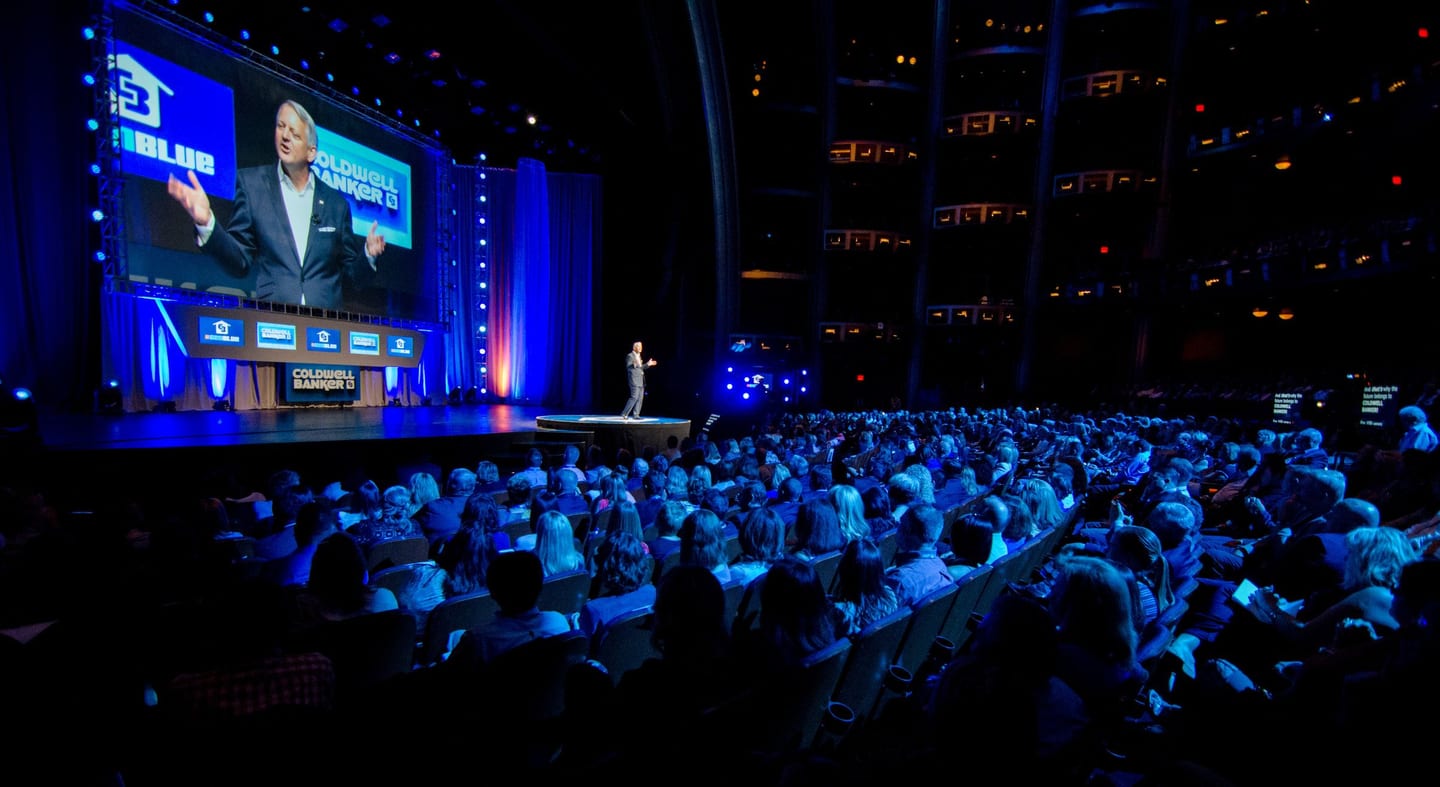 A speaker on stage before a full auditorium under blue stage light at a corporate general-session keynote