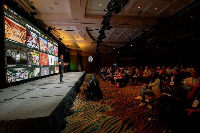A speaker presenting on a long runway stage before a large screen and a seated audience at a conference keynote