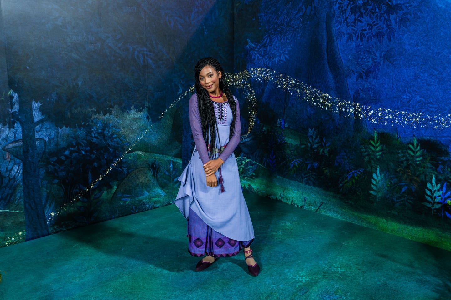 A performer in costume as Asha from Disney’s Wish, in a lavender dress with long braids, standing on a hand-painted storybook forest carpet set as a trail of gold sparkles arcs across the backdrop behind her