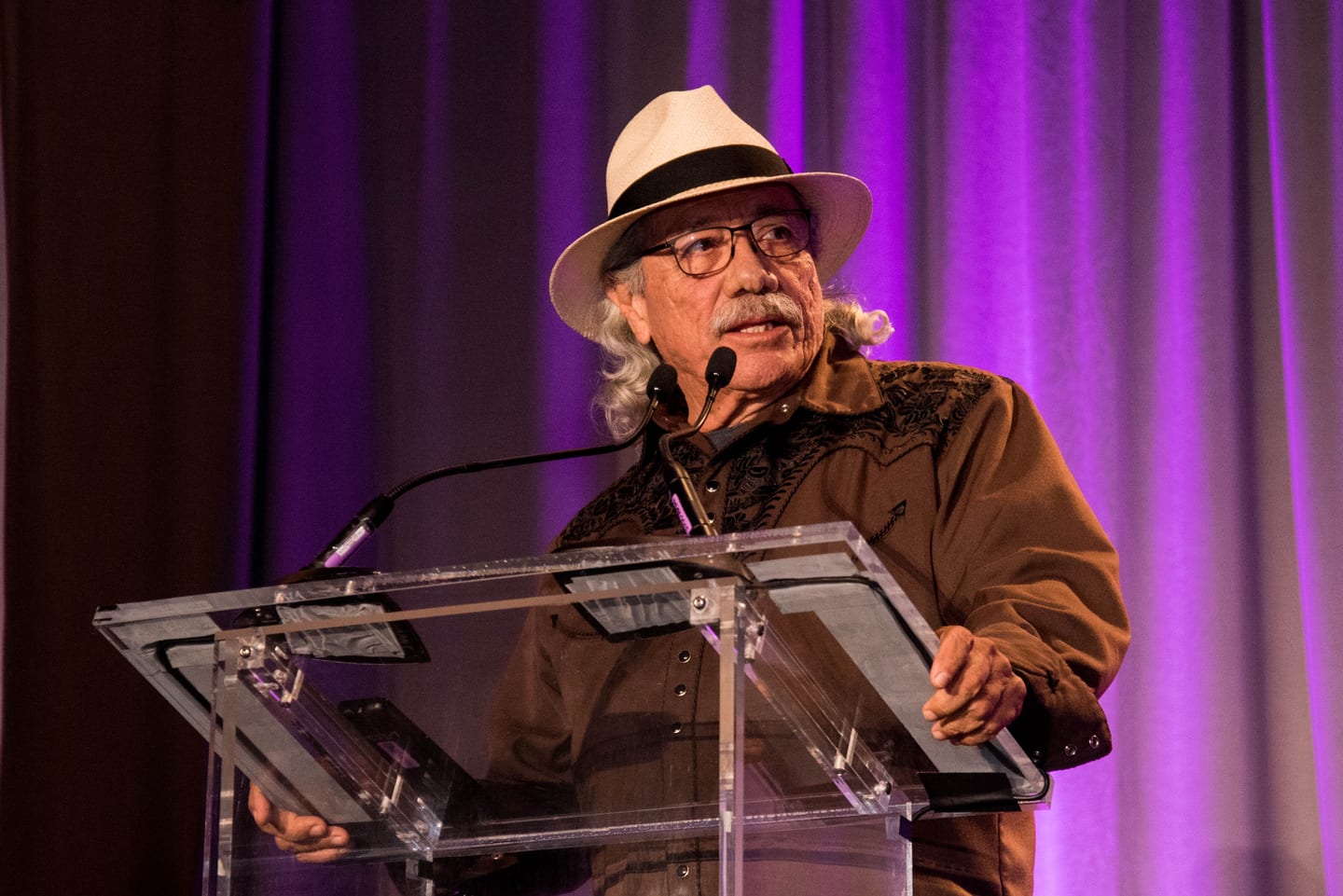 Edward James Olmos in a panama hat and glasses speaking into two microphones at a clear lucite podium, a purple-lit drape behind him