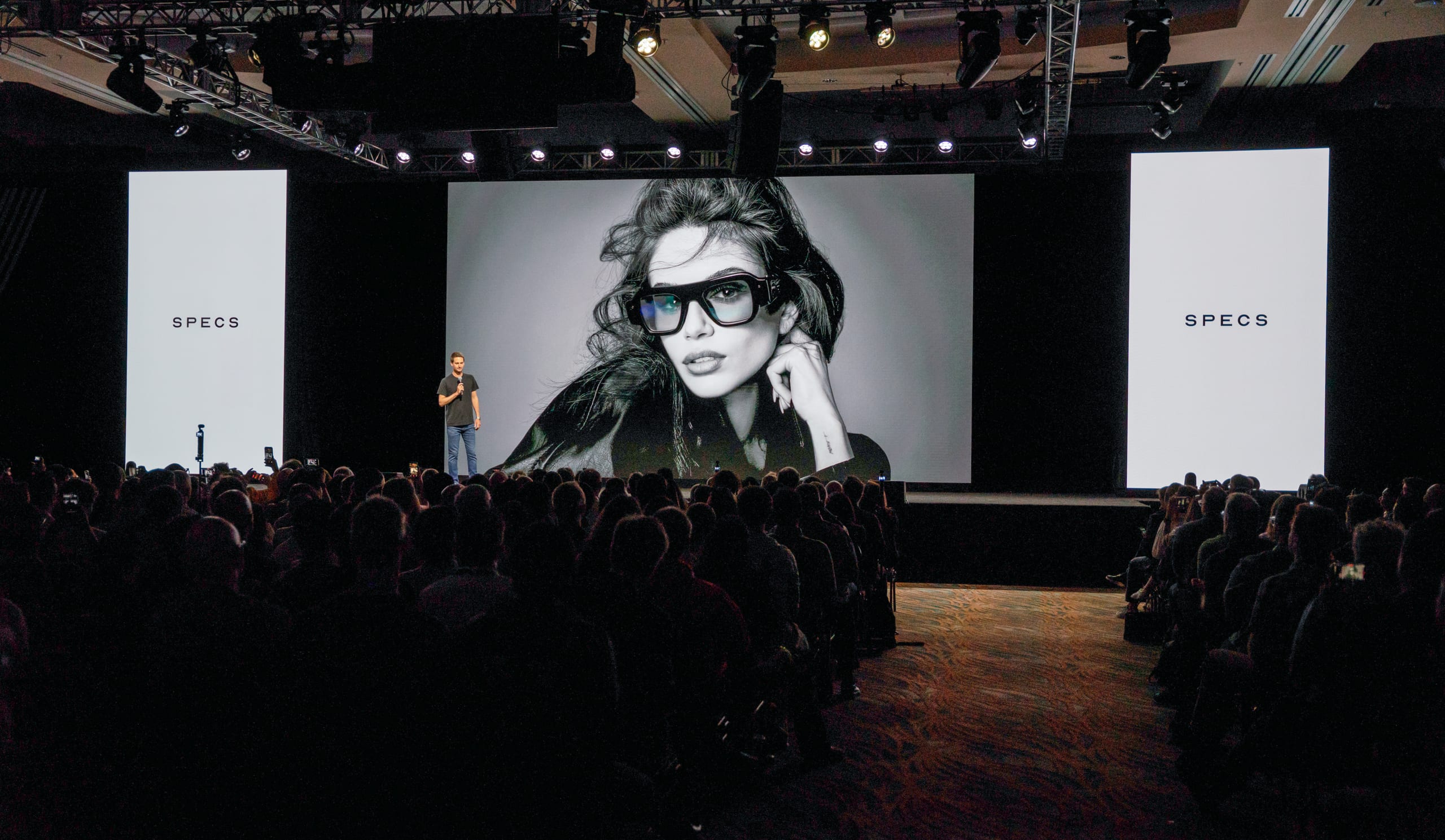 Snap CEO Evan Spiegel alone on a wide stage with a microphone introducing Specs, a large black-and-white campaign portrait of a model wearing the AR glasses on the screen behind him and lit SPECS panels either side, a packed audience silhouetted in the foreground