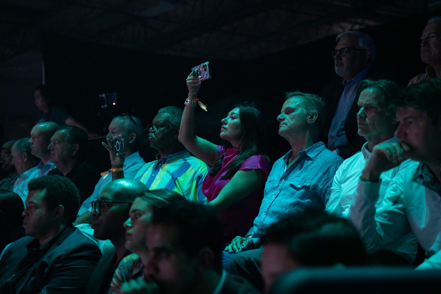 The audience in teal light with phones raised toward the stage