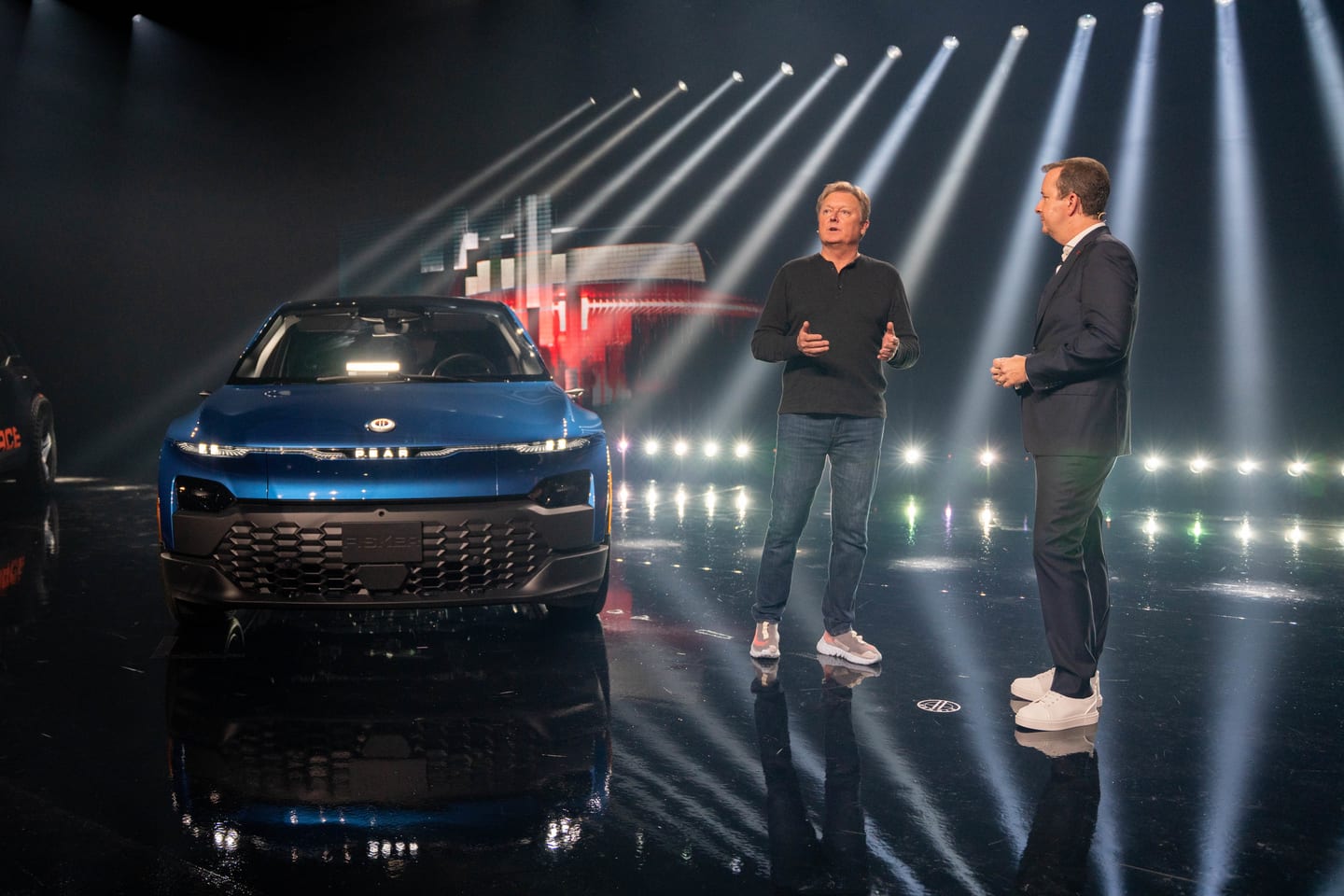 Two presenters on stage beside a blue Fisker SUV under white beams