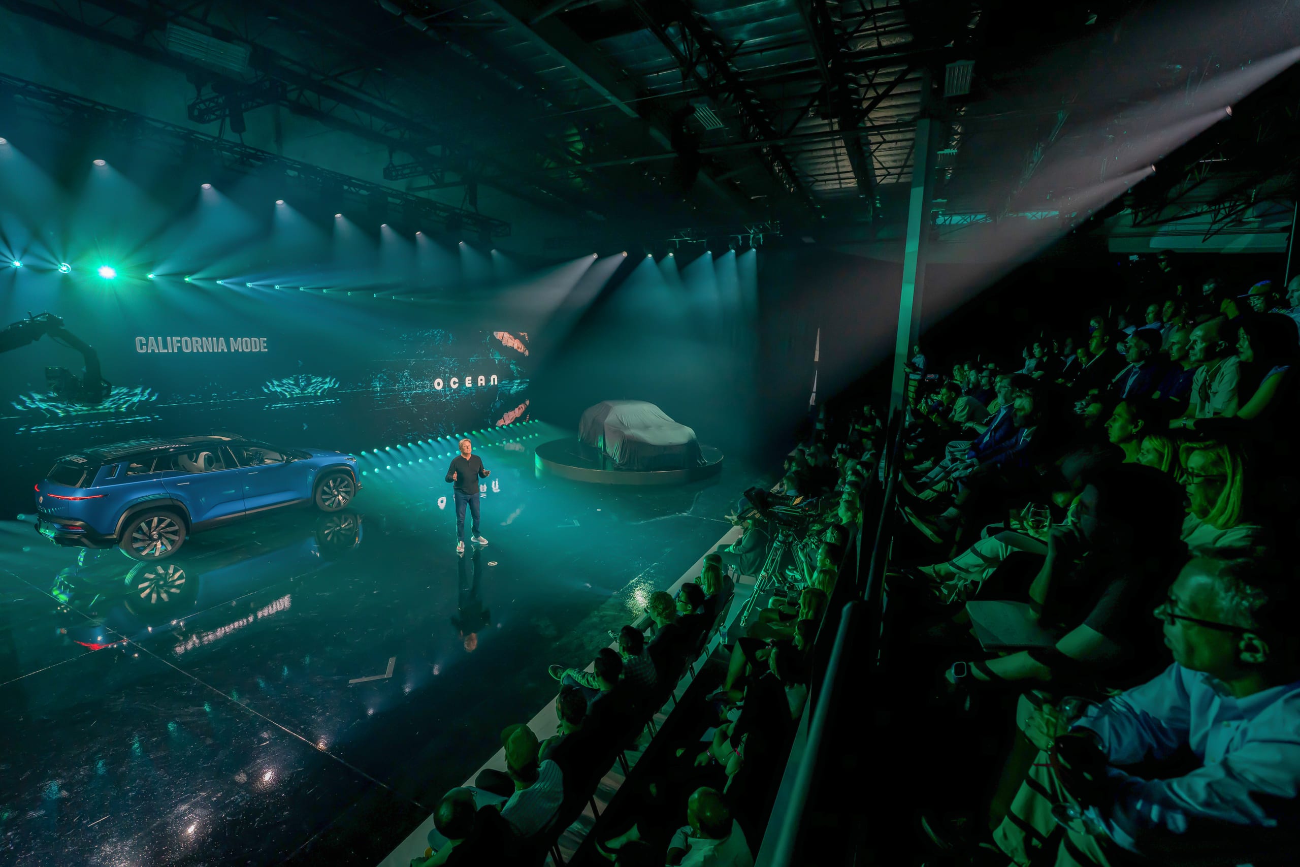 A presenter on a teal-lit stage beside a blue Fisker Ocean and a covered vehicle, facing a full tiered audience at the Fisker product reveal