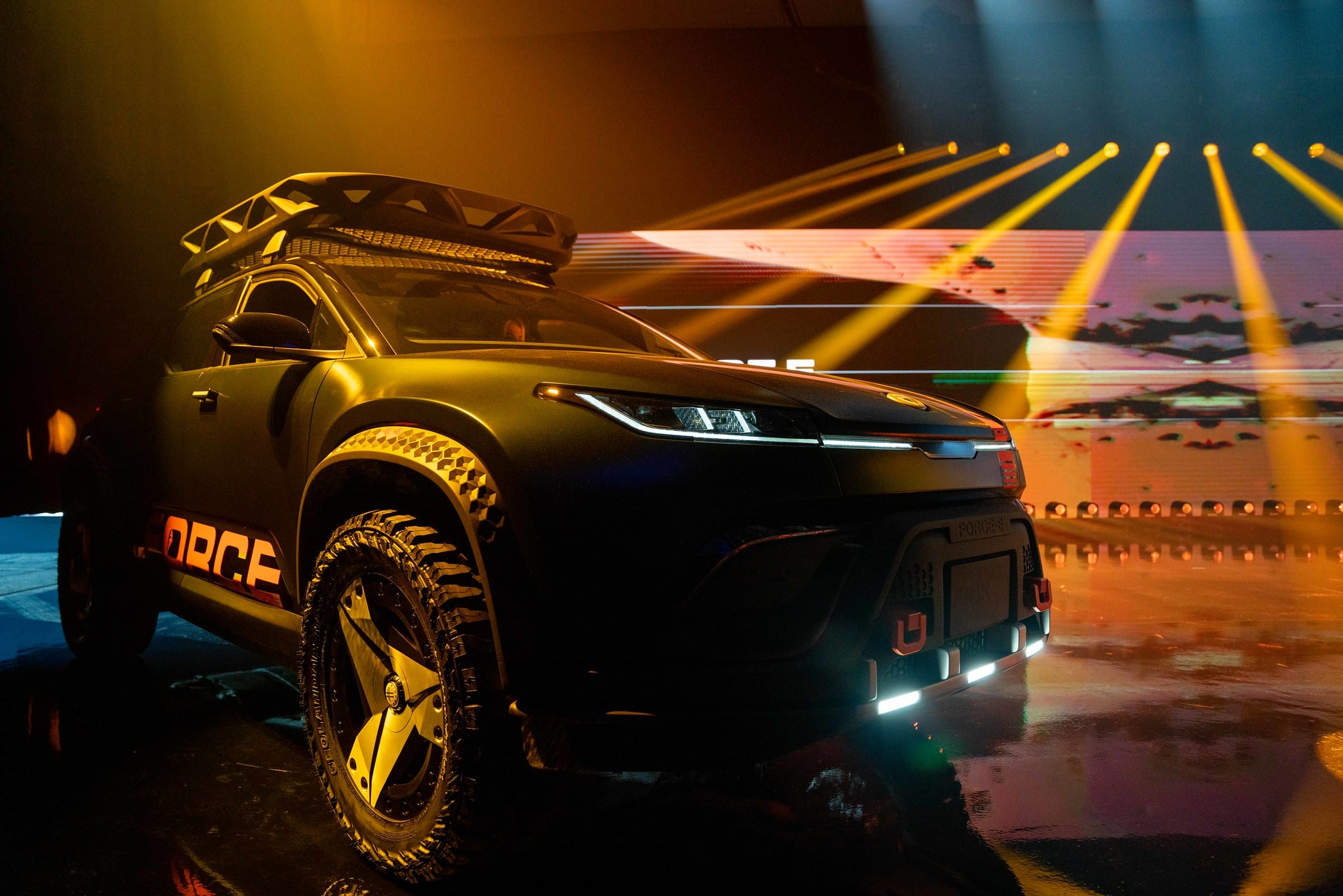 The matte-black Fisker Ocean Force E off-road SUV with roof rack and off-road tires on a reveal stage under dramatic yellow light beams, reflected on a wet floor