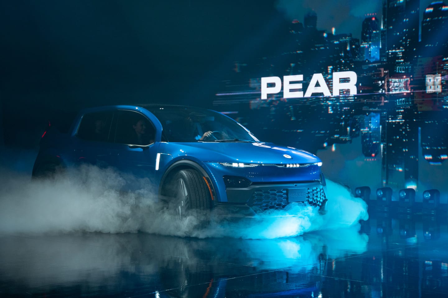 The blue Fisker PEAR emerging through stage fog under a PEAR screen
