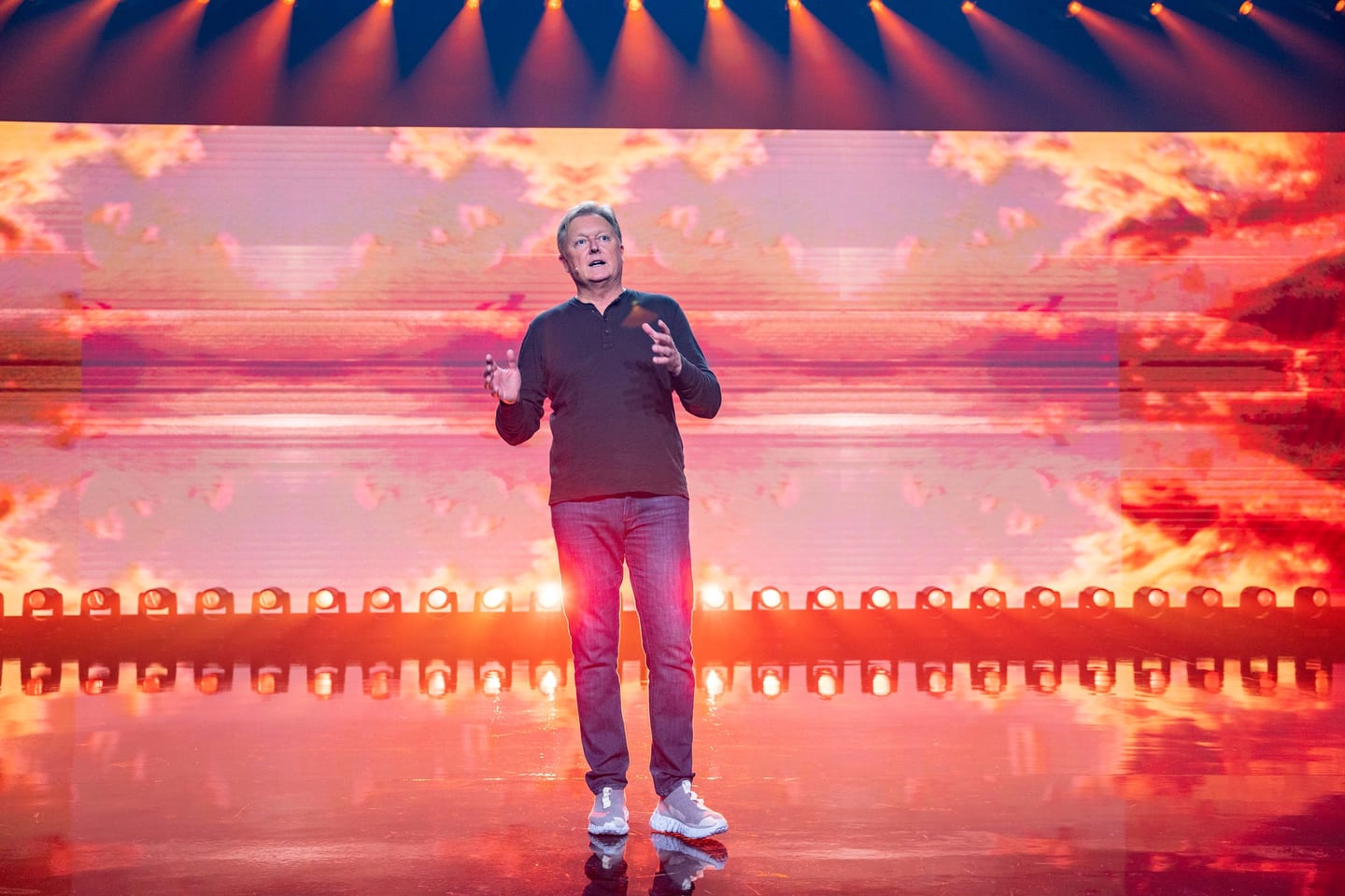 A presenter mid-sentence against a wide orange screen
