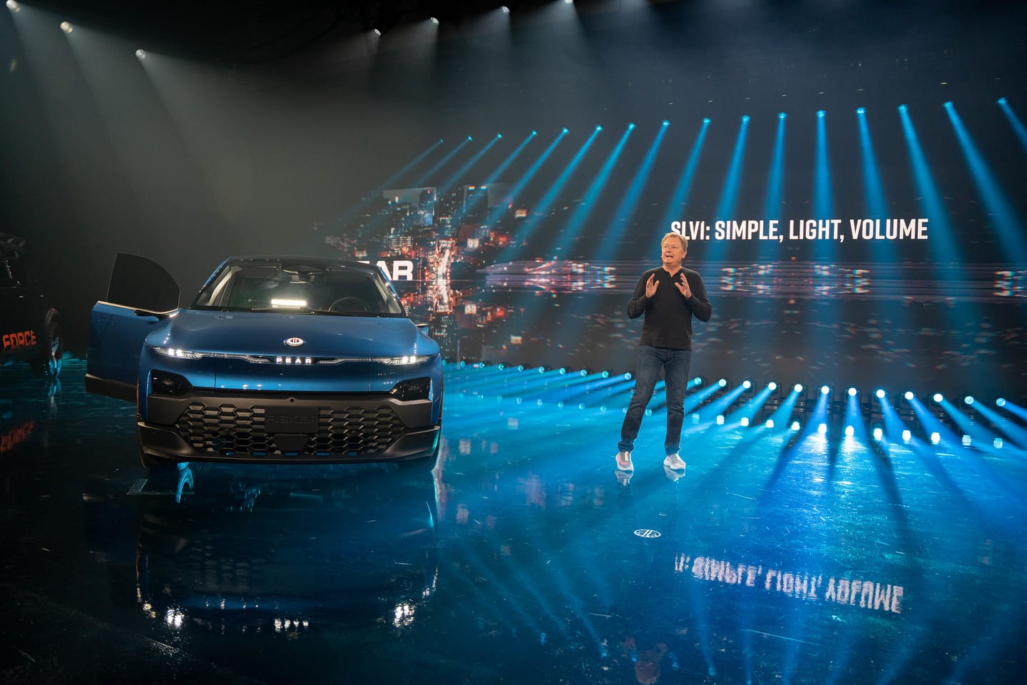 A presenter on stage beside a blue Fisker vehicle, the screen reading SLV: Simple, Light, Volume