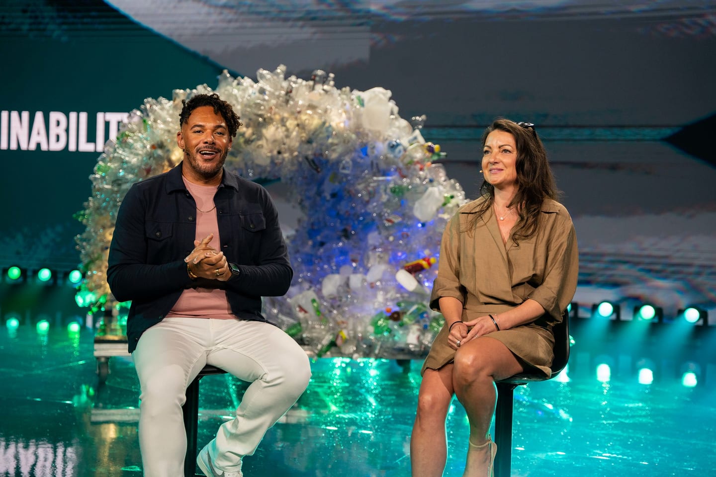 Two speakers seated on stage in front of a recycled-plastic sculpture