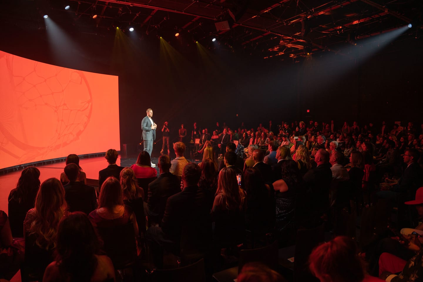 Wide view of the darkened ceremony room, the audience seated facing the lit stage