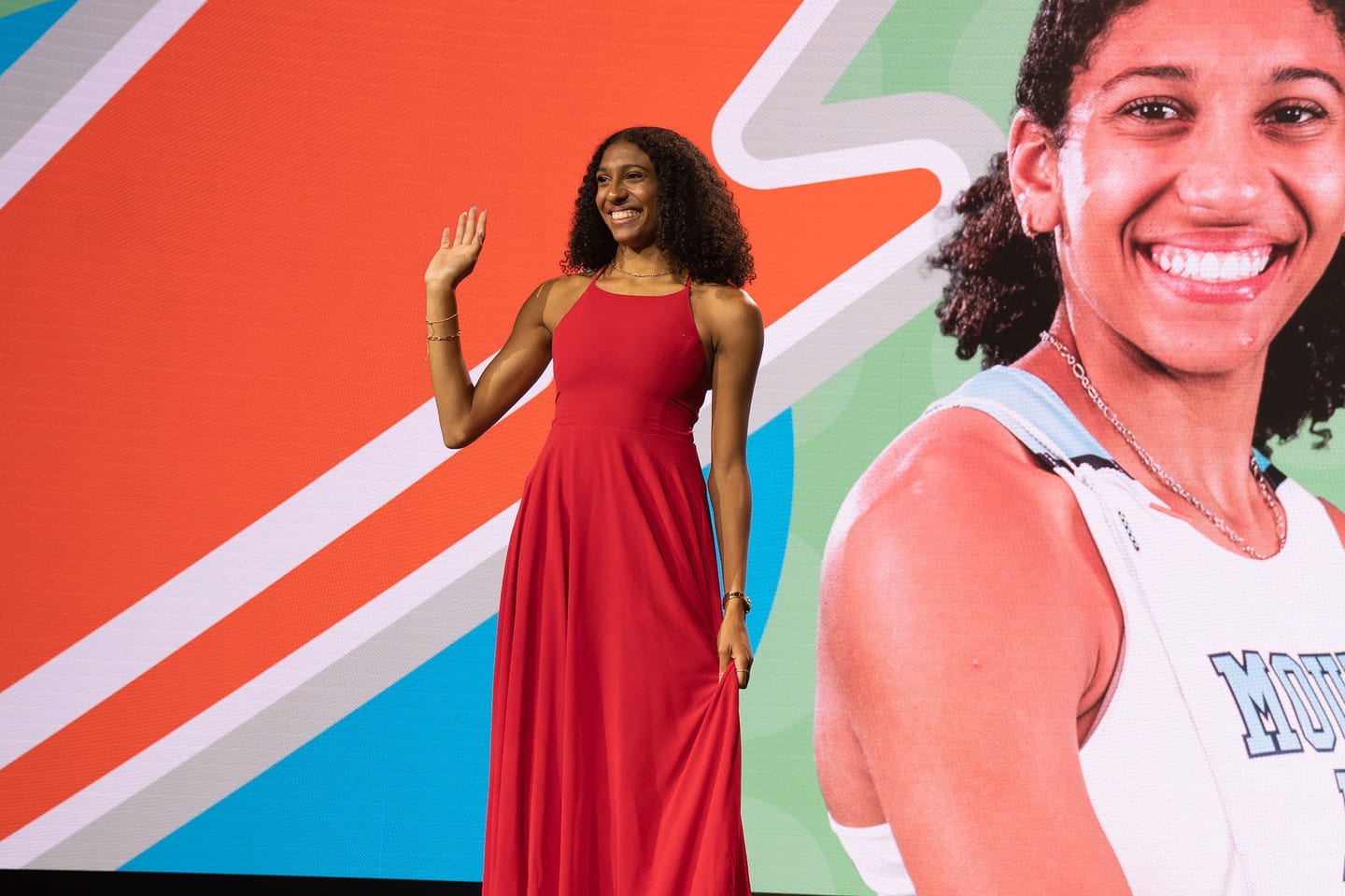 An award winner on stage in a red dress in front of a basketball player graphic