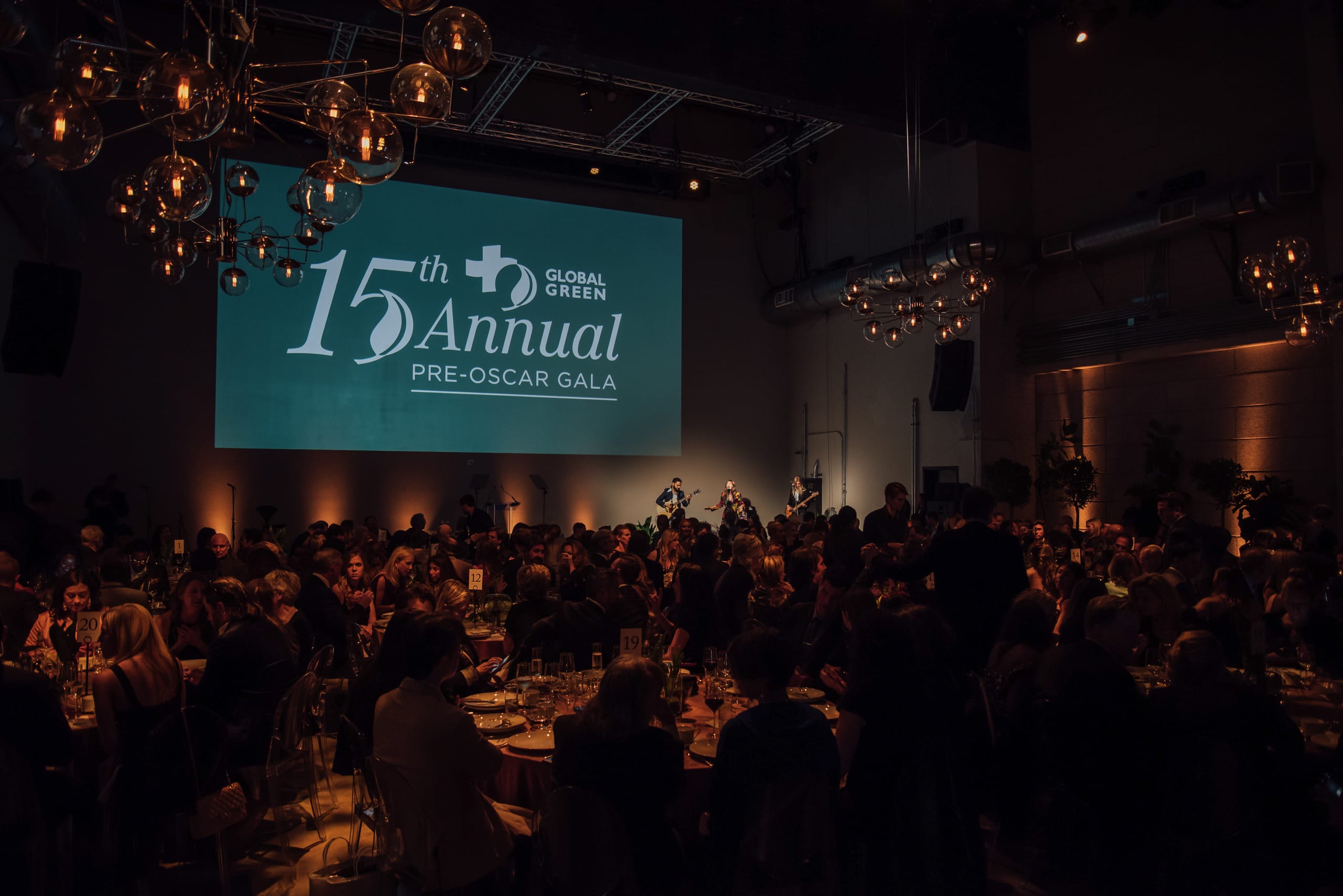 The gala room at NeueHouse Hollywood with guests seated at candlelit tables beneath a screen reading 15th Annual Global Green Pre-Oscar Gala