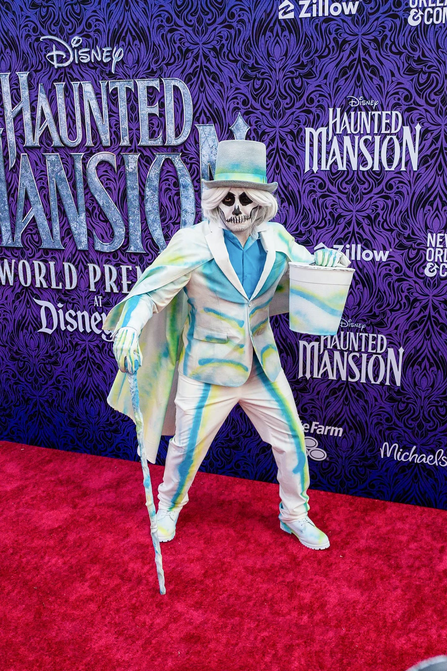 A performer in skull makeup, a grey top hat and a pale tie-dyed suit posed wide-legged on the red carpet holding a cane and a hatbox, against the purple damask Disney Haunted Mansion World Premiere step-and-repeat