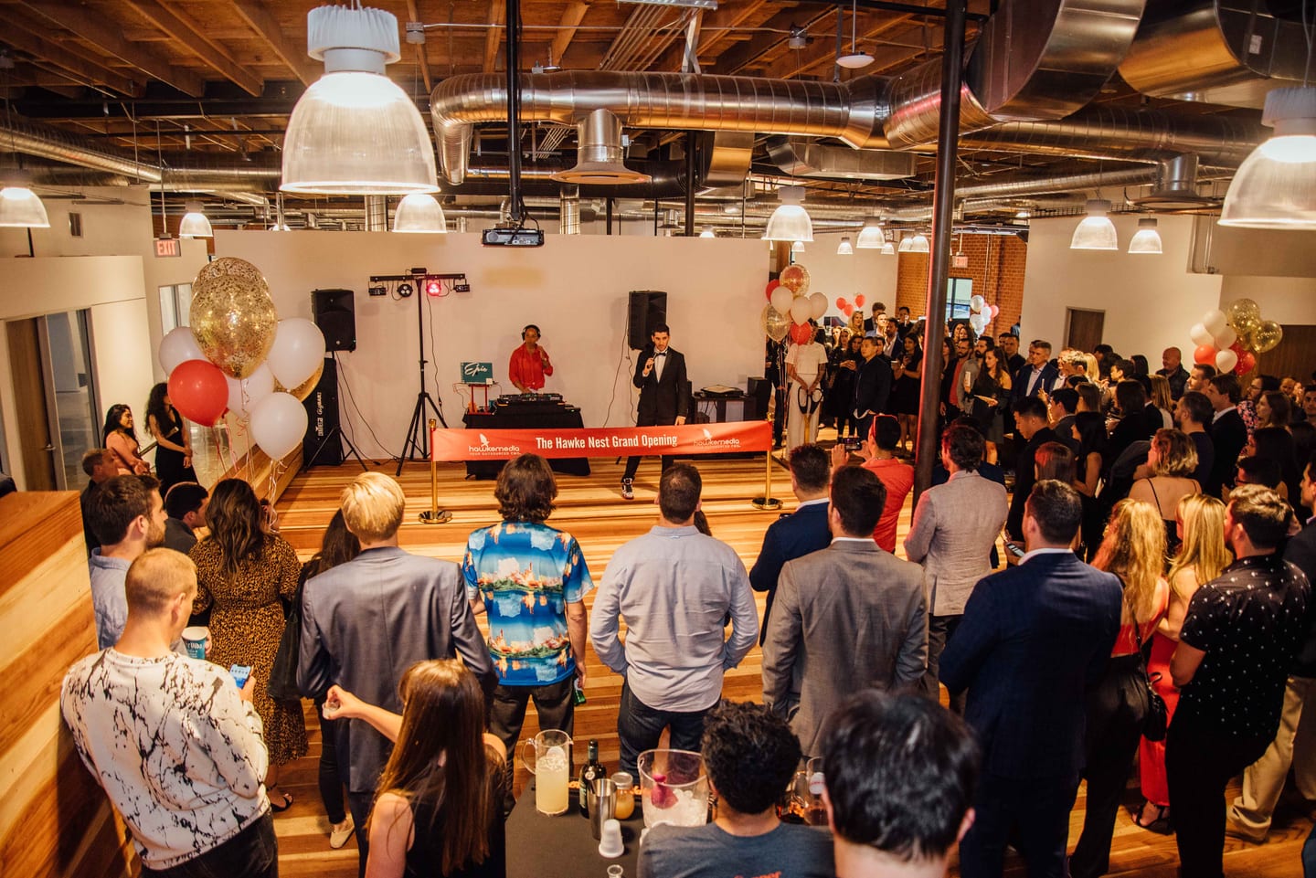 A packed room of guests watching a host with a microphone behind a ribbon reading The Hawke Nest Grand Opening, a DJ and balloon clusters on the platform in a exposed-beam office loft