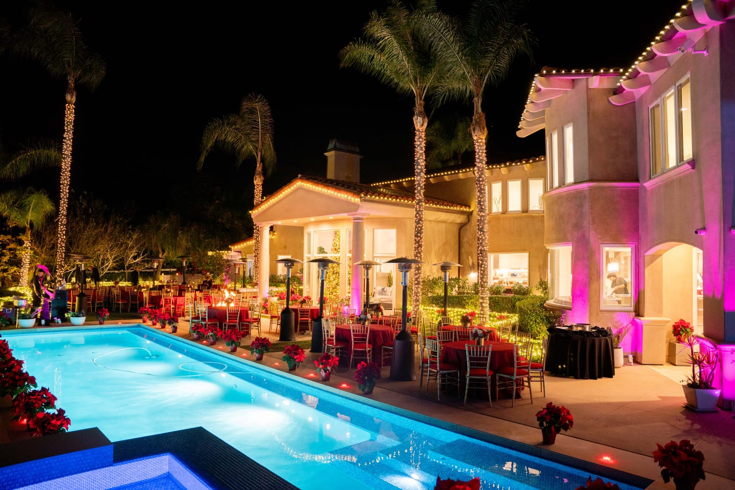 A private estate decorated for a holiday party at night, with string lights in the palms, a glowing pool, magenta uplighting and dinner tables dressed in red with poinsettias
