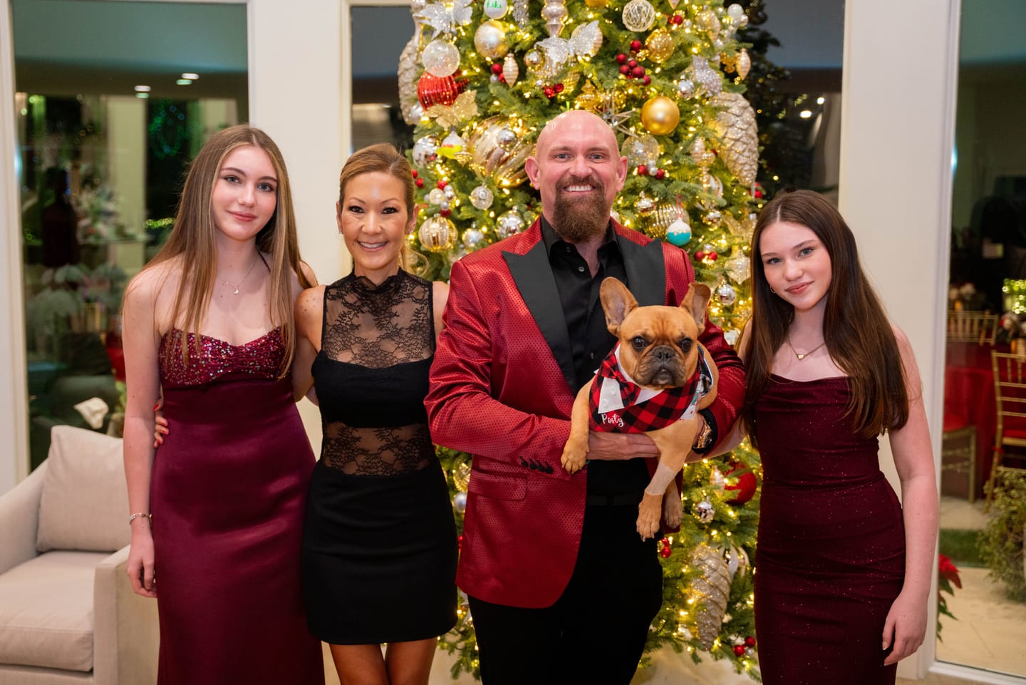 A family in holiday formalwear posed with their French bulldog in front of a decorated Christmas tree at a private holiday party