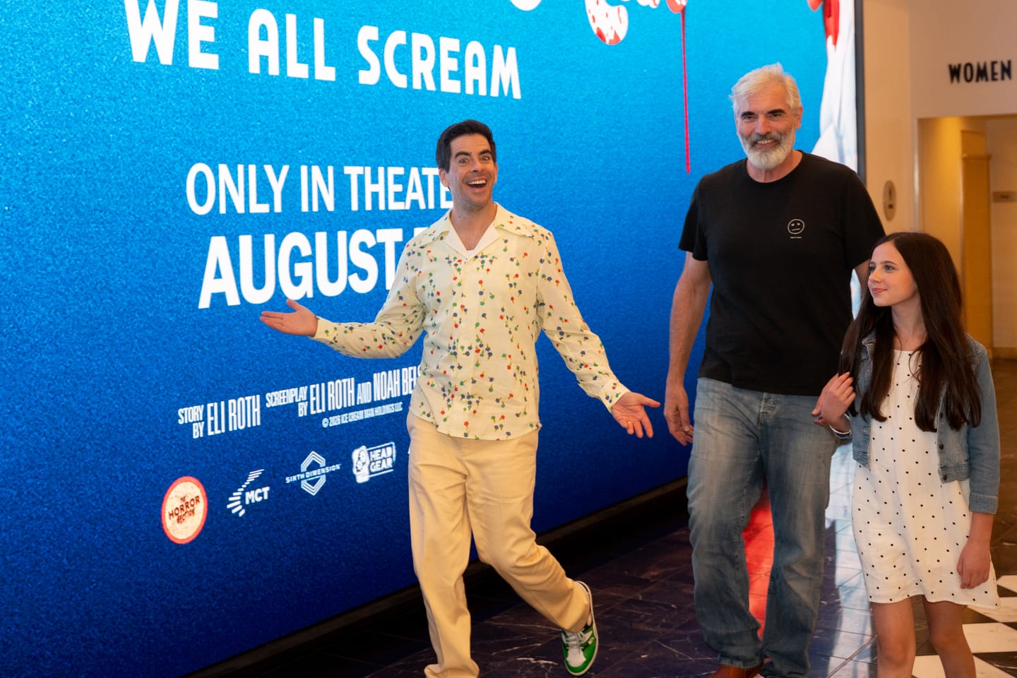 Eli Roth walking into the theatre with his arms out, passing a wall-sized lit display reading We All Scream, Only In Theaters August 7th